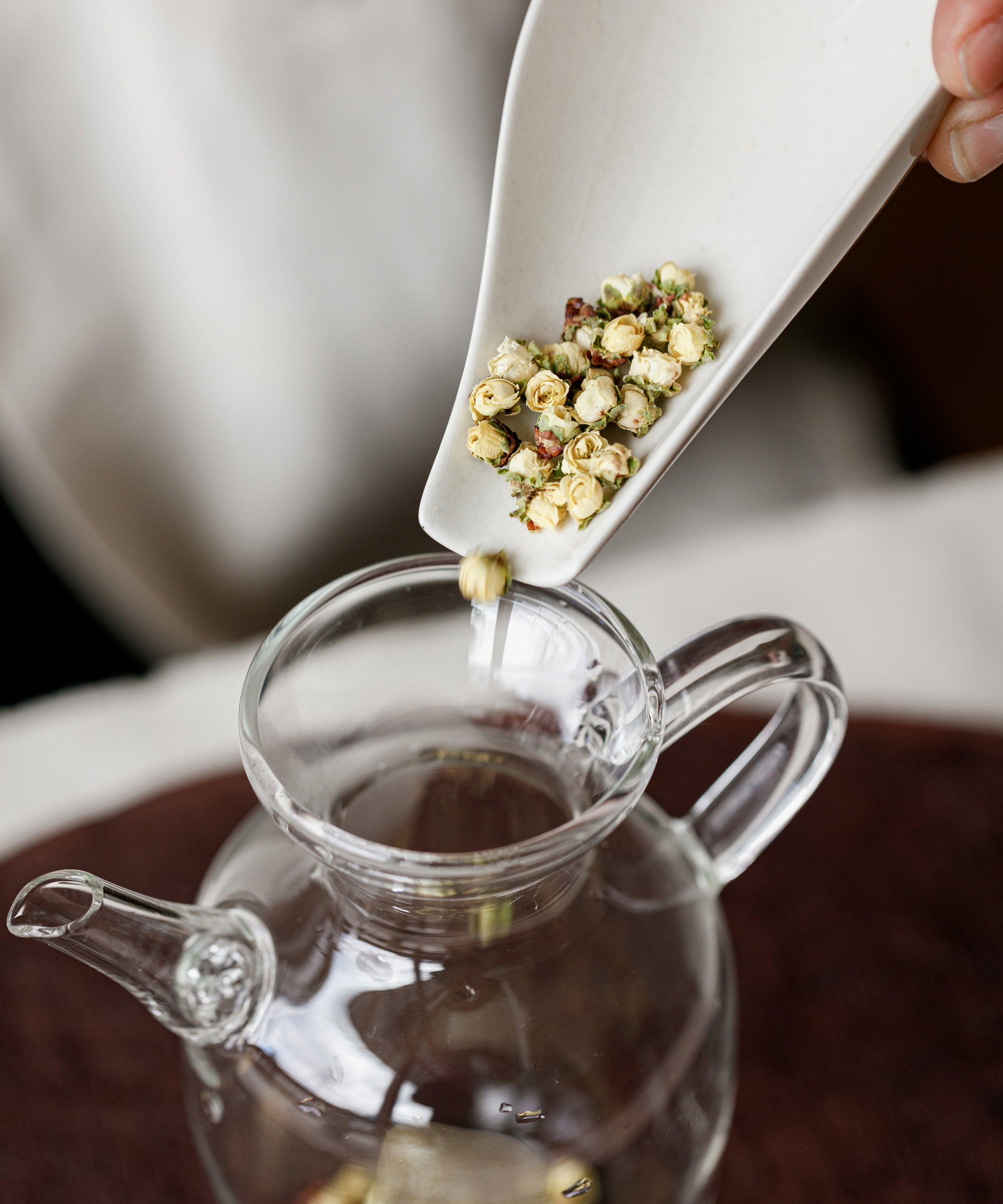 Hand scooping dried plum blossoms with a measure into a glass teapot